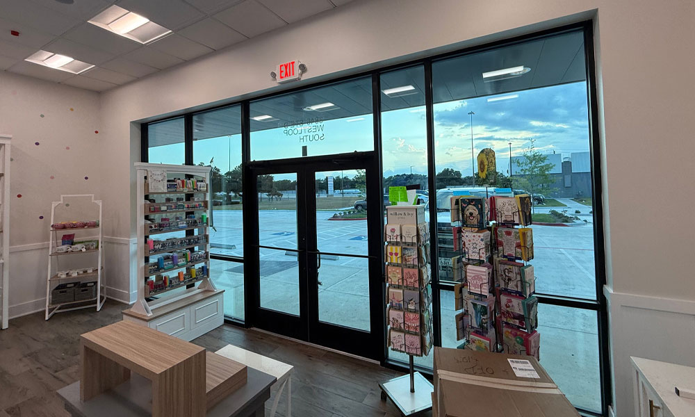 Interior view of a Nothing Bundt Cakes location showcasing a welcoming bakery space with display shelves for products, a wooden table, and large glass doors leading outside, emphasizing the retail environment's design and layout.