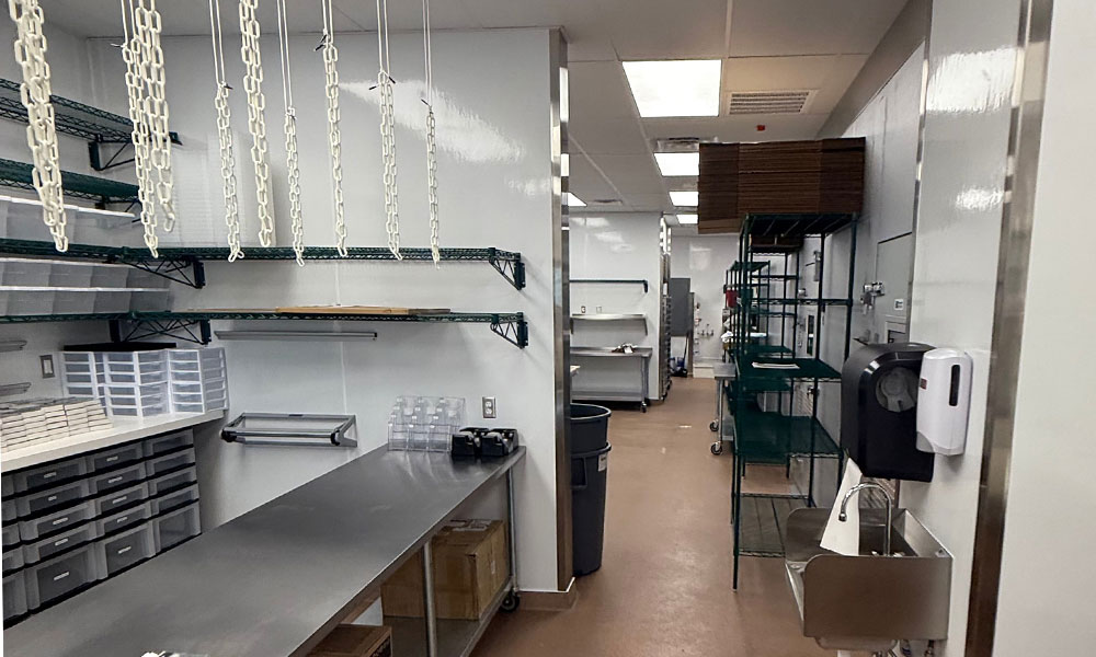 Commercial bakery kitchen interior with stainless steel counters, shelving units, storage bins, and food preparation areas designed for Nothing Bundt Cakes operations.