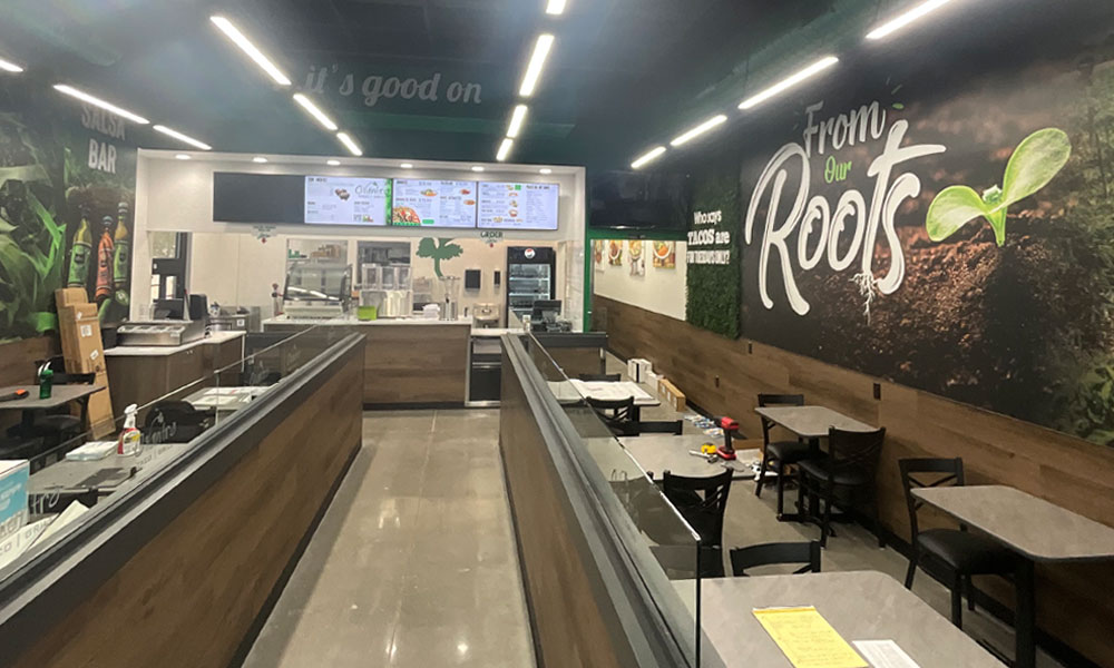 Interior of Cilantros Taco Bar in Celina, TX, featuring modern design elements, a salsa bar, menu displays, and vibrant wall art with the phrase "From Our Roots," highlighting the fast-casual dining atmosphere.