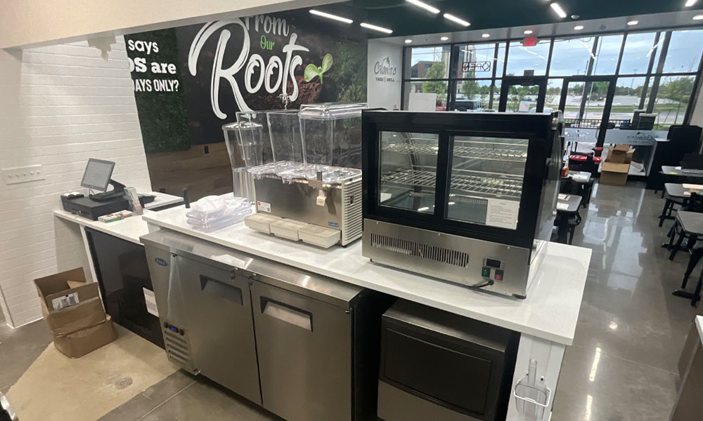 Modern taco bar counter with kitchen equipment, display cases, and a digital order station, featuring the Cilantros brand logo and a "From Our Roots" mural in a vibrant fast-casual dining setting.