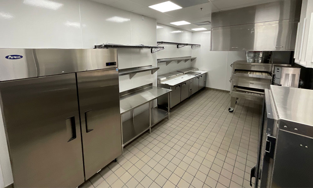 Commercial kitchen interior featuring stainless steel appliances, including a refrigerator and prep tables, designed for Mountain Mike's Pizza tenant build-out by Kinetic Builders Corp.