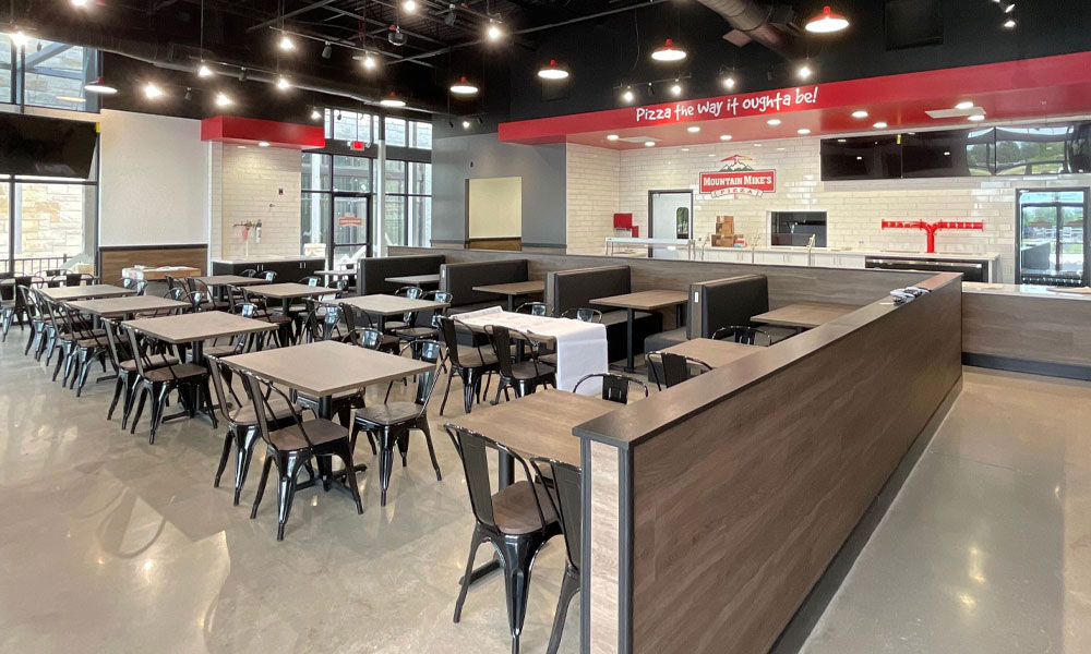 Interior of a Mountain Mike’s Pizza restaurant featuring modern seating arrangements, black metal chairs, and a welcoming atmosphere, with a visible service counter displaying the brand logo and the slogan "Pizza the way it oughta be!"