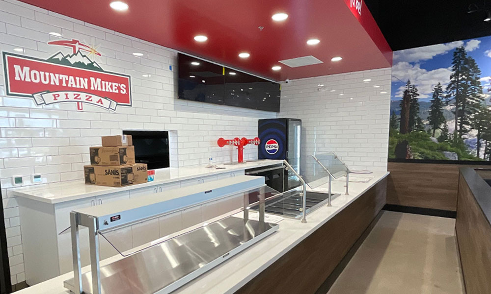 Mountain Mike's Pizza restaurant interior featuring logo, counter space with Pepsi machine, and a scenic mural of trees, illustrating a family-friendly dining environment.