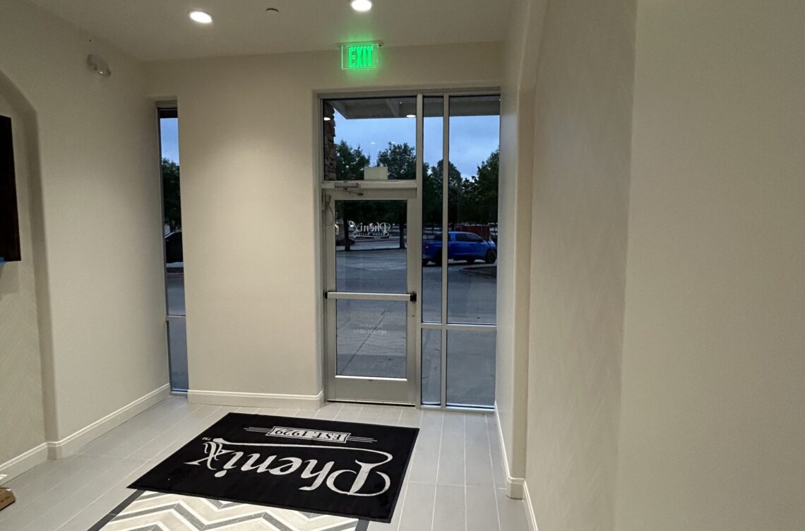 Interior view of a commercial space entrance featuring a black welcome mat with "Established 1990" text, glass doors leading outside, and a green exit sign, showcasing Kinetic Builders Corp.'s craftsmanship in commercial construction.