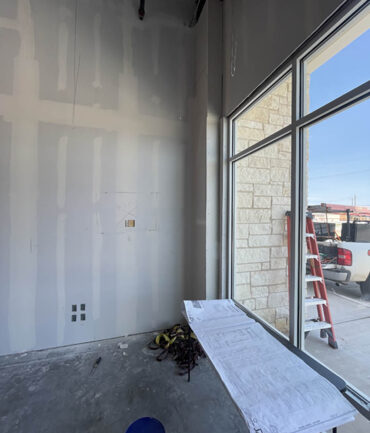 Interior construction site showing drywall installation, construction plans on a table, and a ladder near a large window, reflecting Kinetic Builders Corp.'s commercial project work.
