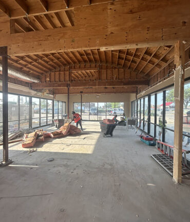 Interior view of a commercial construction site with exposed wooden beams, workers preparing the space, and large windows allowing natural light, showcasing Kinetic Builders Corp.'s craftsmanship in progress.