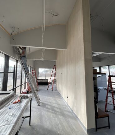 Construction worker on ladder installing fixtures in commercial space with unfinished walls and visible scaffolding, showcasing Kinetic Builders Corp's commitment to quality in restaurant build-outs.
