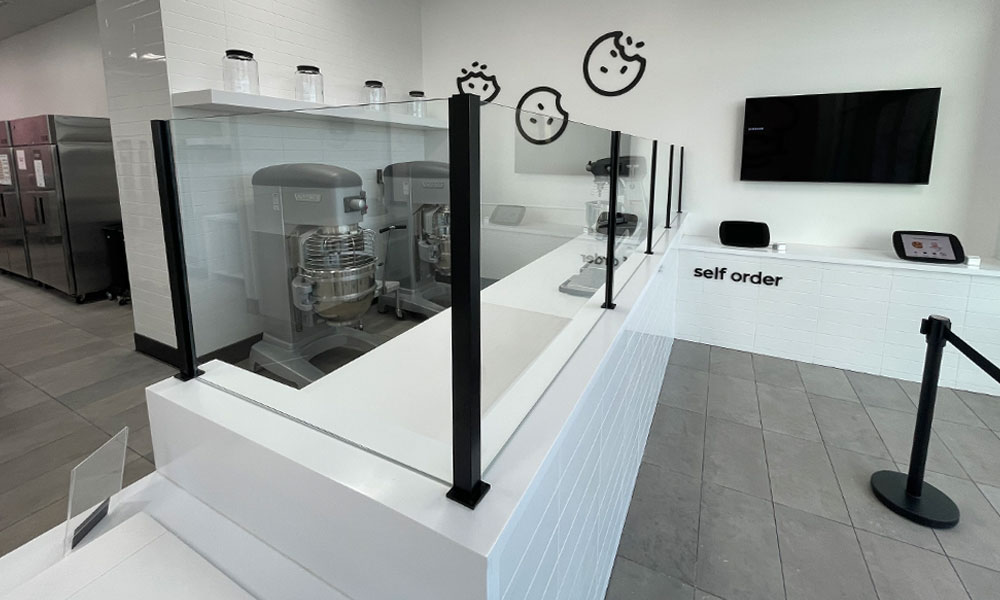 Modern Crumbl Cookies self-order area featuring glass barriers, kitchen equipment, and branding elements, designed for efficient customer interaction and operational functionality.