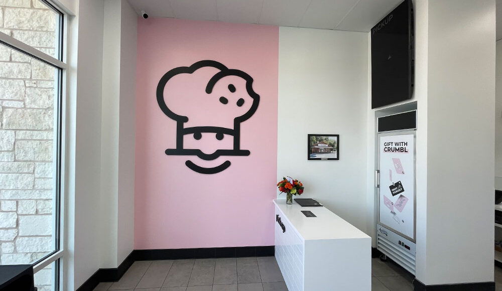 Crumbl Cookies store interior featuring a large chef logo on a pink wall, modern white counter, and promotional signage for gifts, highlighting Kinetic Builders Corp's tenant build-out expertise.