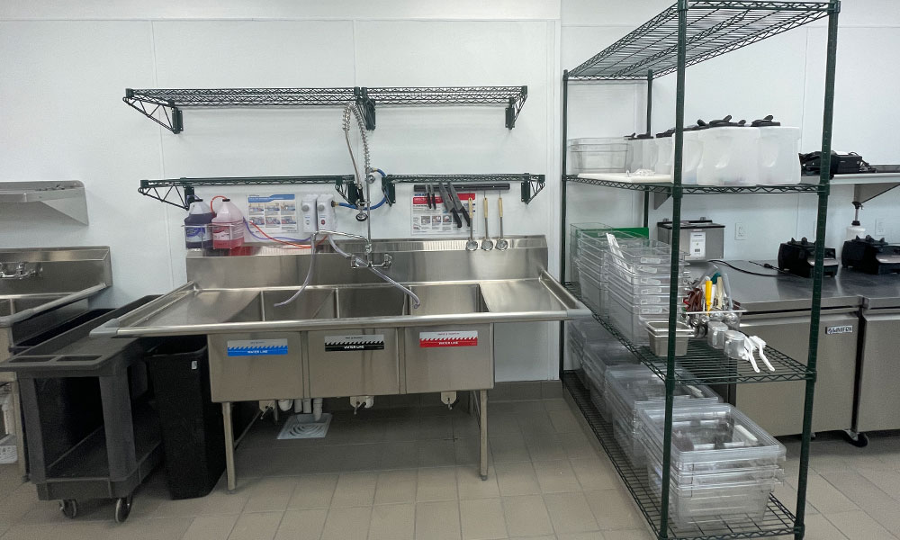 Commercial kitchen workspace featuring stainless steel sinks, shelving units, and organized storage containers, illustrating efficient design for Nekter Juice Bar's food retail tenant build-out by Kinetic Builders Corp.