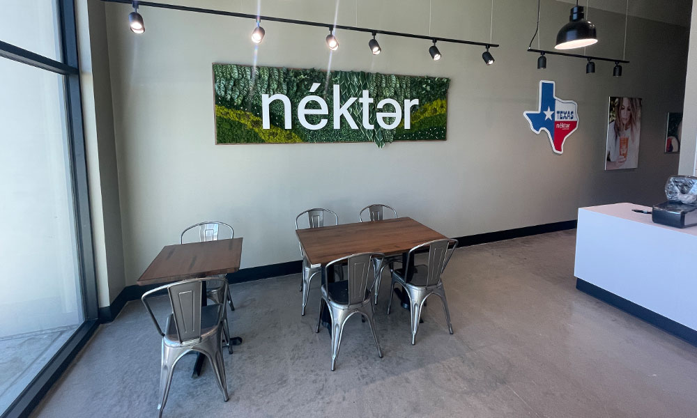 Interior of a Nekter Juice Bar location in Texas, featuring a large green wall sign with the brand name "néktər," modern metal tables and chairs, and a clean, minimalist design emphasizing wellness and functionality.