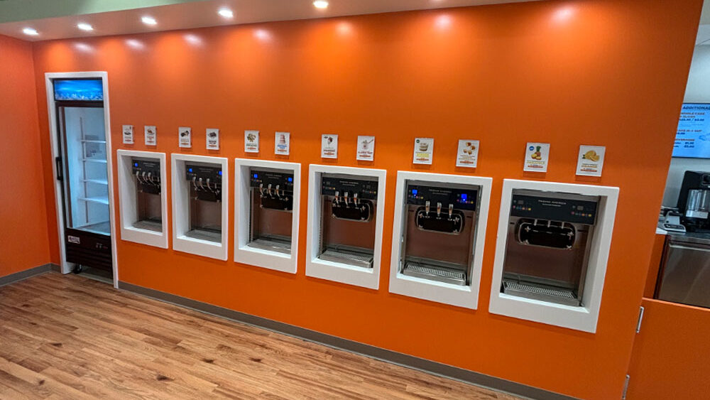 Self-serve frozen yogurt machines against an orange wall, showcasing various flavor options with signage above each machine, emphasizing efficiency in restaurant build-out design.