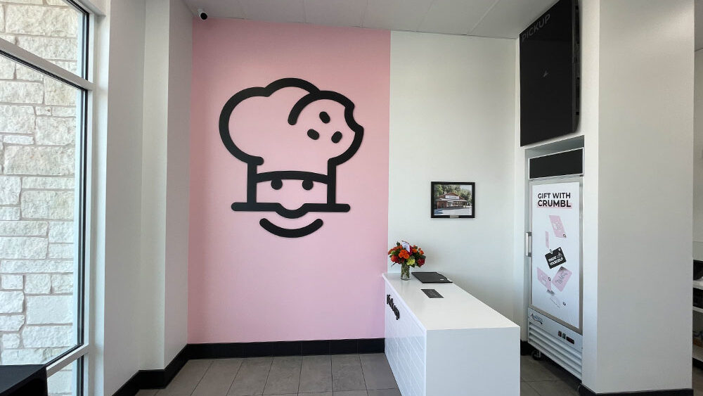Chef cartoon logo on pink wall in modern restaurant entrance, featuring a smiling face and chef's hat, with white service counter and decorative flowers, highlighting inviting atmosphere for dining area renovations.