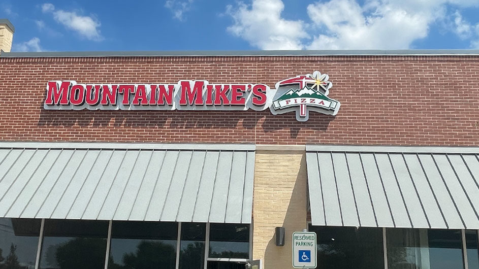 Mountain Mike's Pizza storefront with prominent signage, featuring a red and green logo against a brick background, showcasing the restaurant's name and a mountain graphic, under a blue sky with clouds.